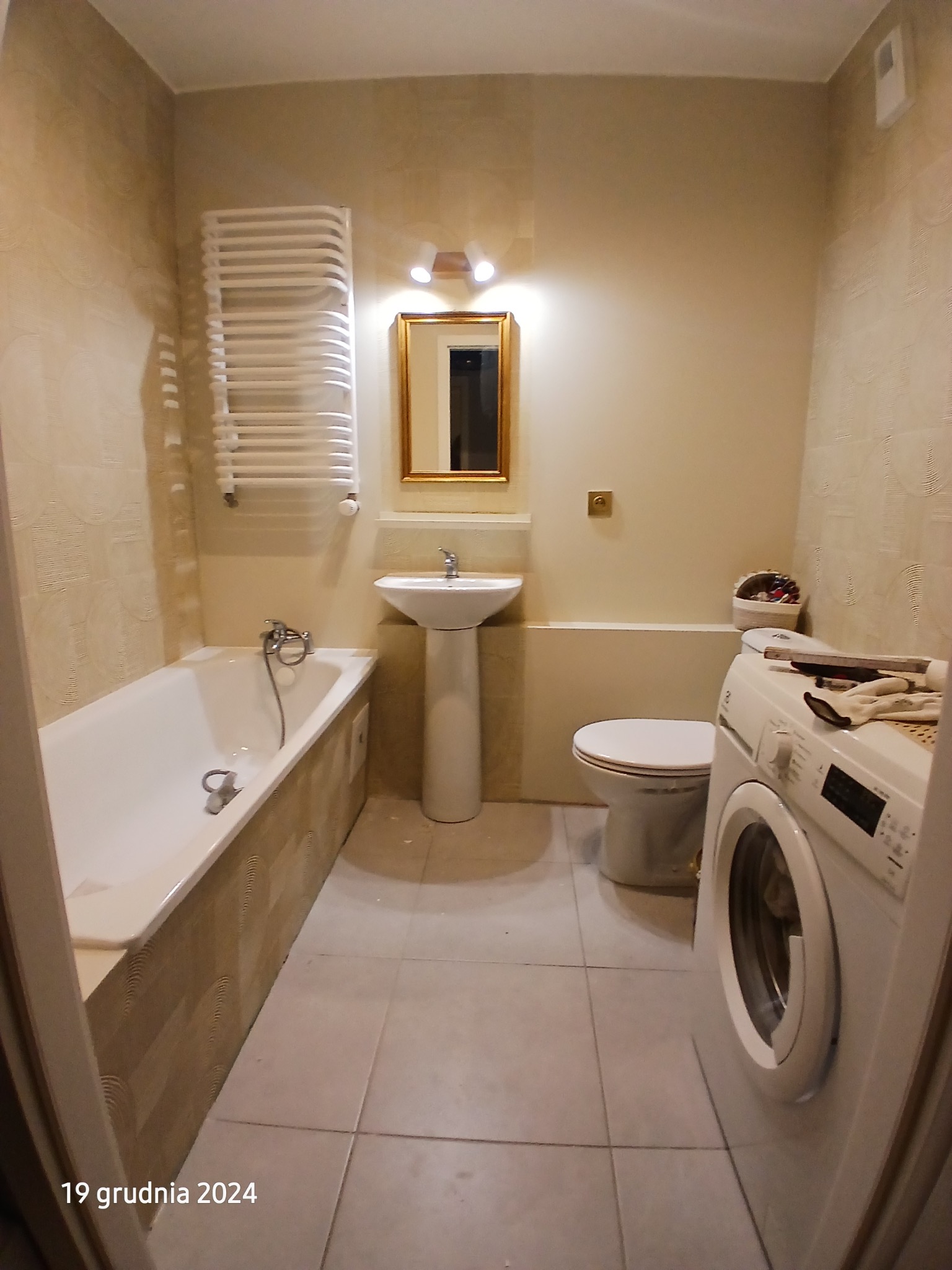 Jasna łazienka po remoncie: wanna, umywalka na postumencie, toaleta, pralka. Beżowe płytki na ścianach i podłodze. Grzejnik drabinkowy i lustro w złotej ramie.