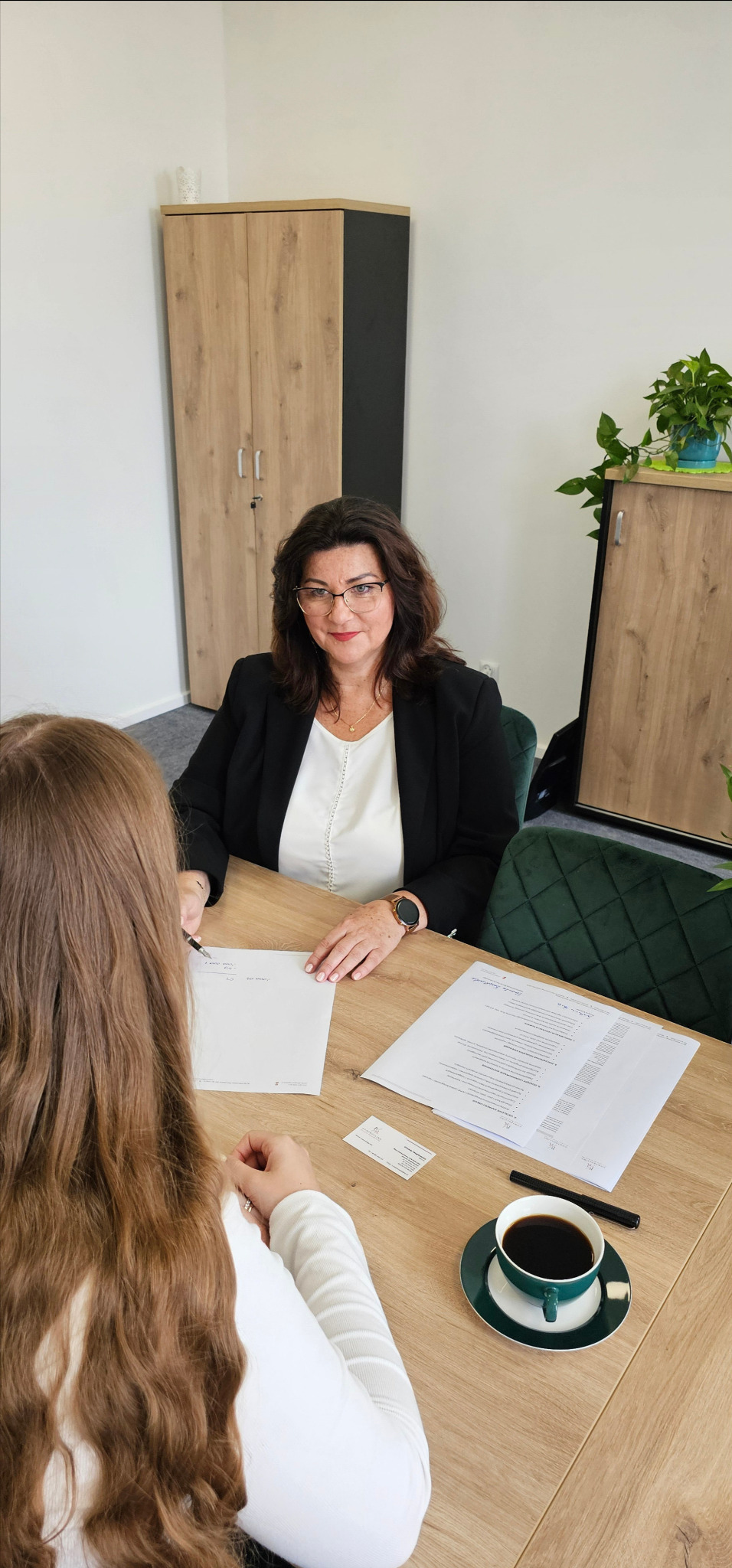 Spotkanie z doradcą: kobieta w okularach omawia dokumenty z klientką przy biurku, kawa, wizytówka, nowoczesne biuro.