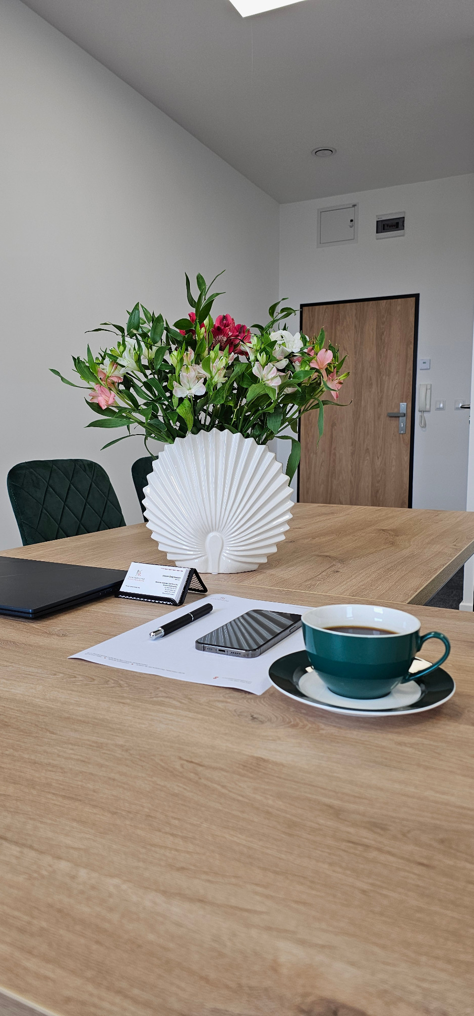 Biuro rachunkowe: drewniany stół z kwiatami, wizytówką, telefonem, długopisem, dokumentem i filiżanką kawy w eleganckim wnętrzu.