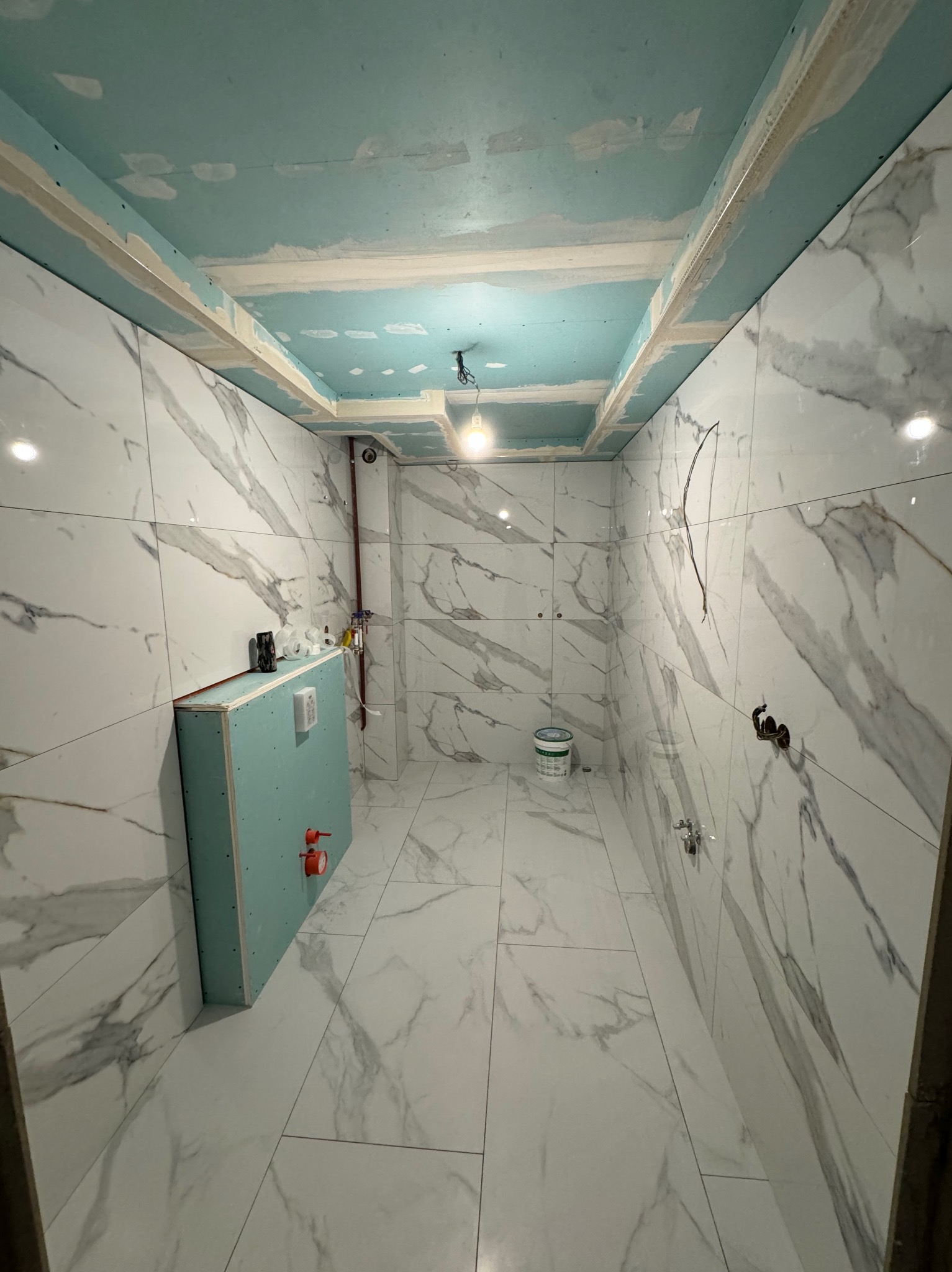 Łazienka w trakcie remontu: białe płytki imitujące marmur na ścianach i podłodze, zabudowa stelaża WC z zielonej płyty karton-gips, sufit z płyt karton-gips przygotowany do malowania, widoczne...