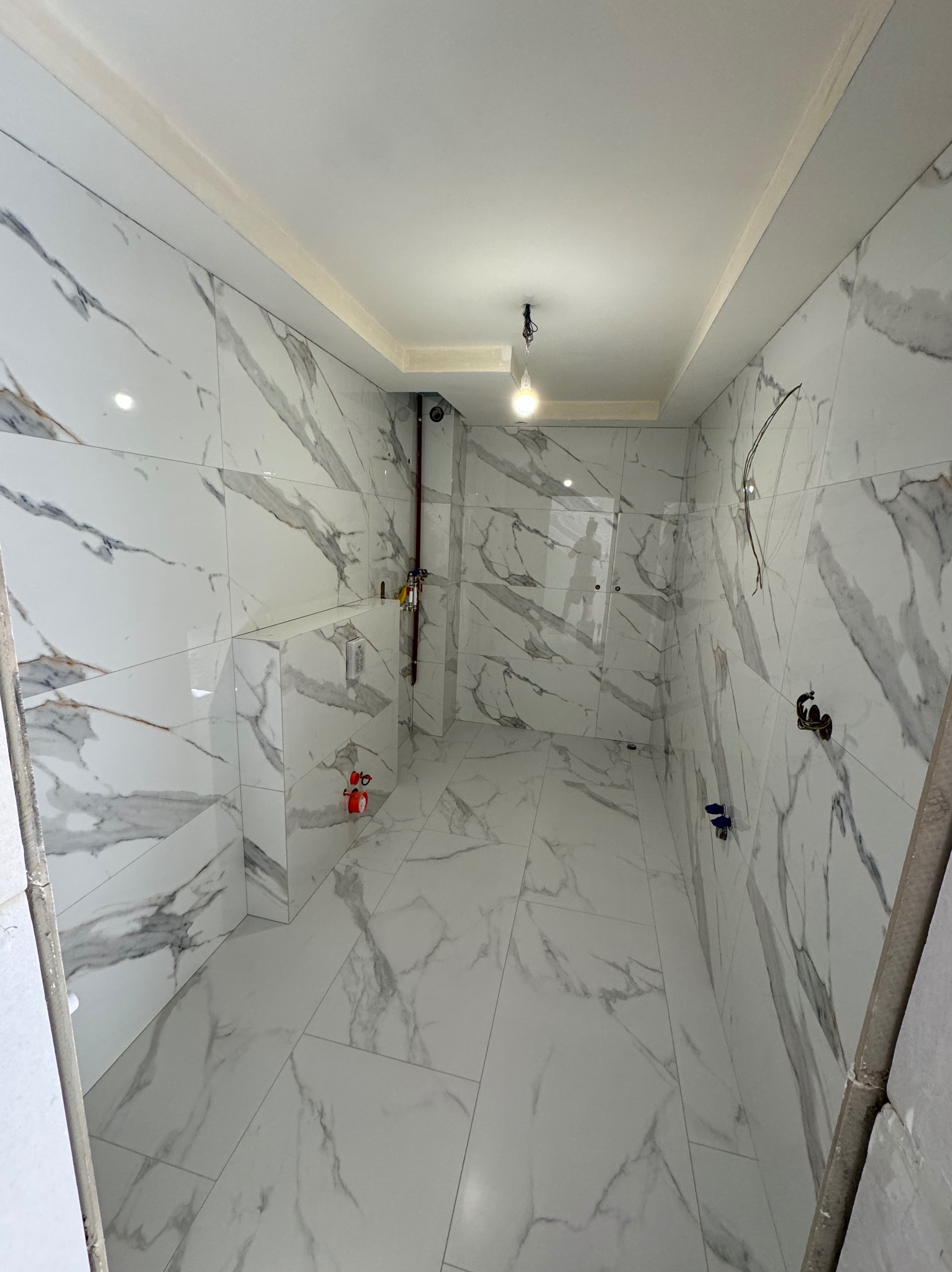 Wnętrze łazienki w trakcie wykańczania, widoczne płytki imitujące marmur na ścianach i podłodze, instalacja wodna i elektryczna w trakcie montażu, sufit z oświetleniem punktowym.