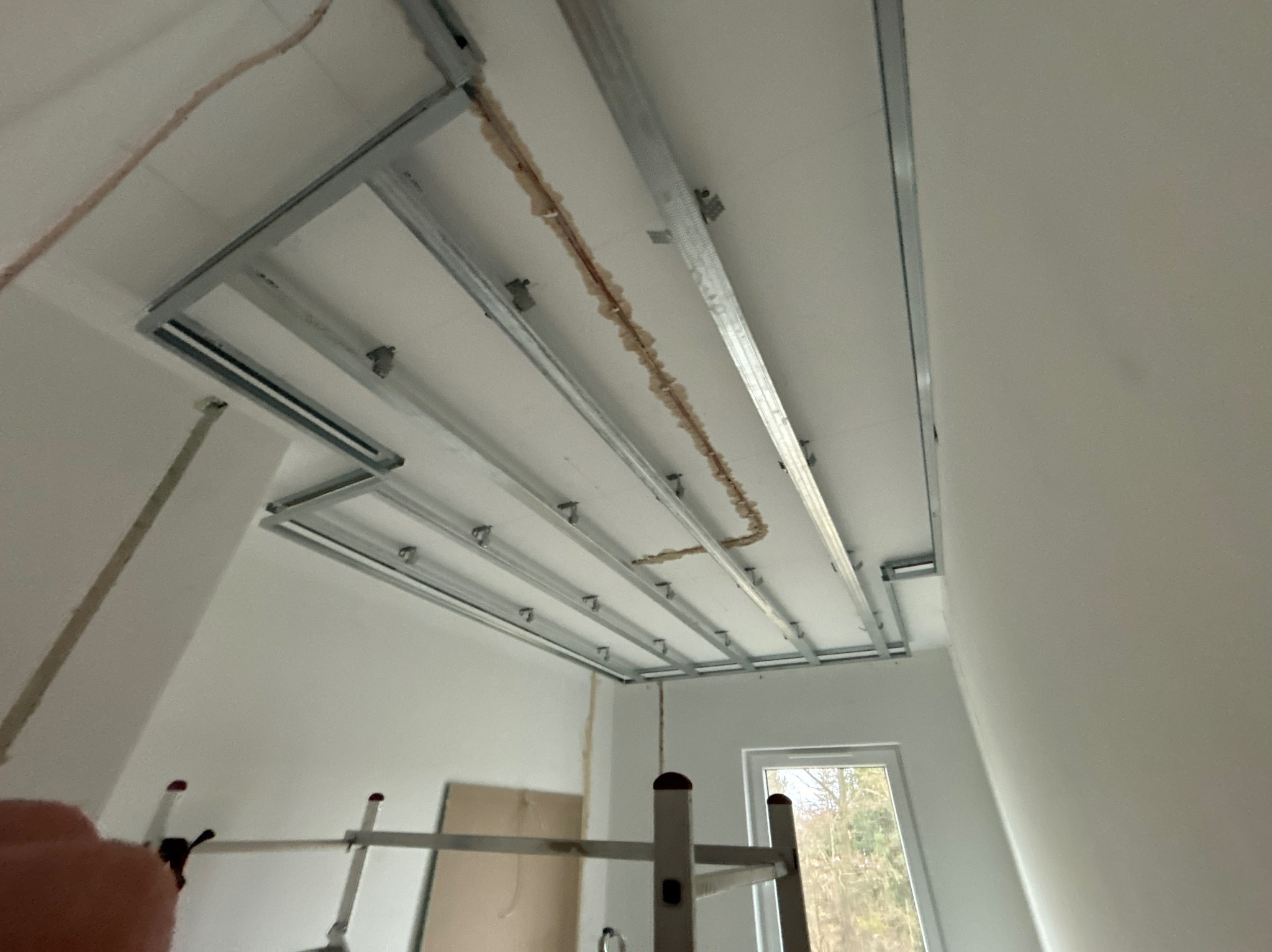 Stelaż sufitu podwieszanego z płyt karton gips, widoczne profile metalowe i częściowo wypełnione przestrzenie, ślady po instalacji elektrycznej na suficie, drabina w tle, okno z widokiem na drzewa.