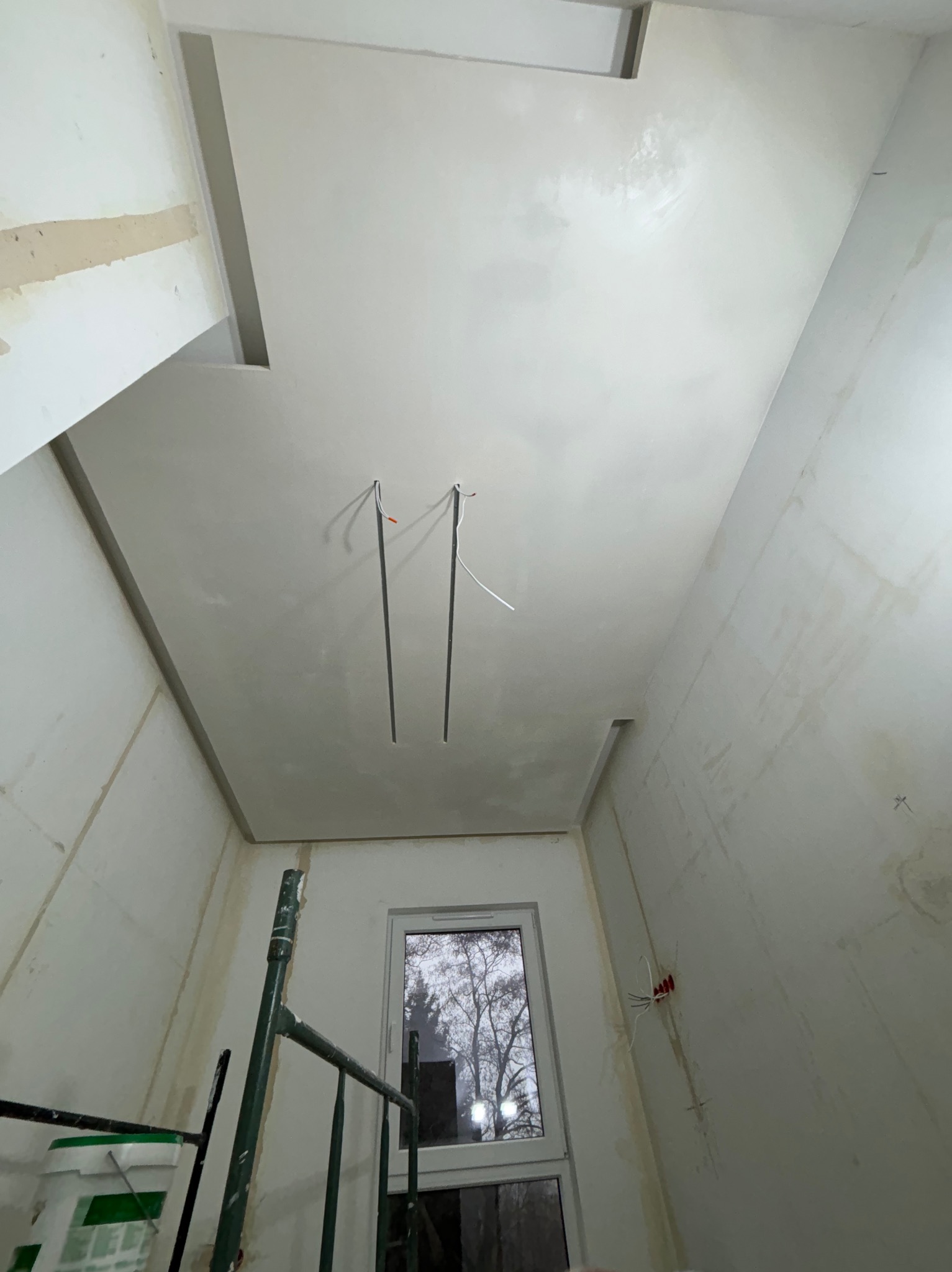 Surowy stan wnętrza w trakcie prac: widoczne ślady po szpachlowaniu na ścianach i suficie, wystające kable z sufitu, okno z widokiem na drzewa, fragment rusztowania z wiadrem farby w rogu.