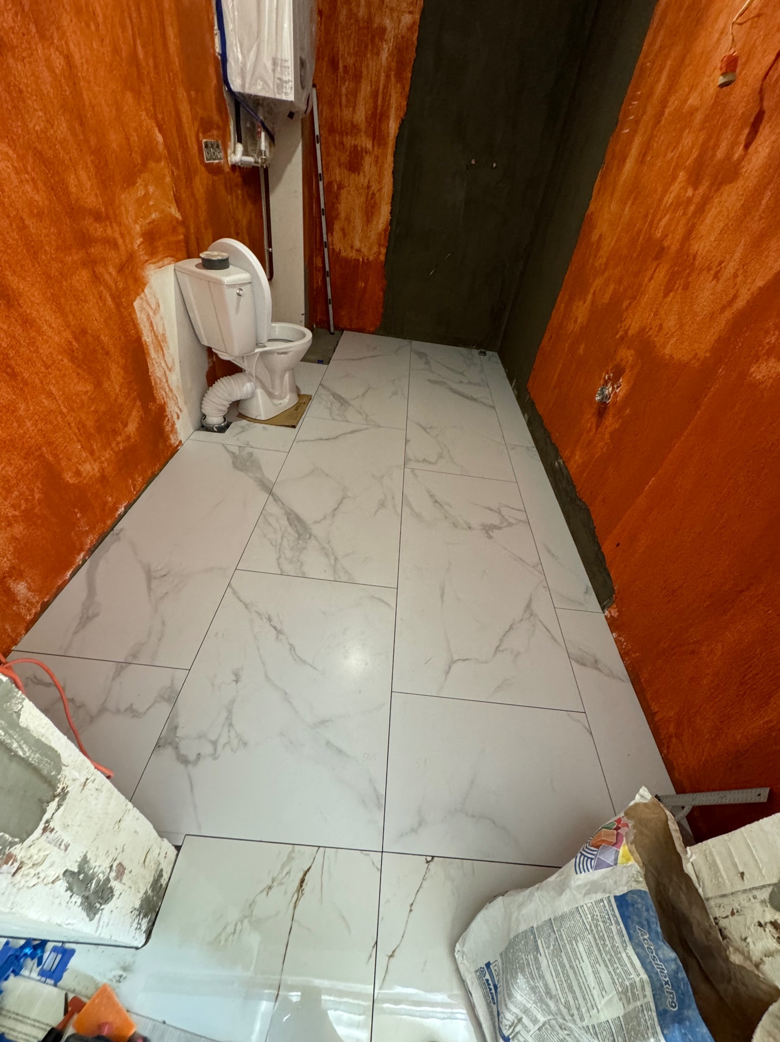 Łazienka w trakcie prac: ułożone białe płytki imitujące marmur na podłodze, pomarańczowe ściany w surowym wykończeniu, widoczny fragment toalety i opakowanie po kleju do płytek.