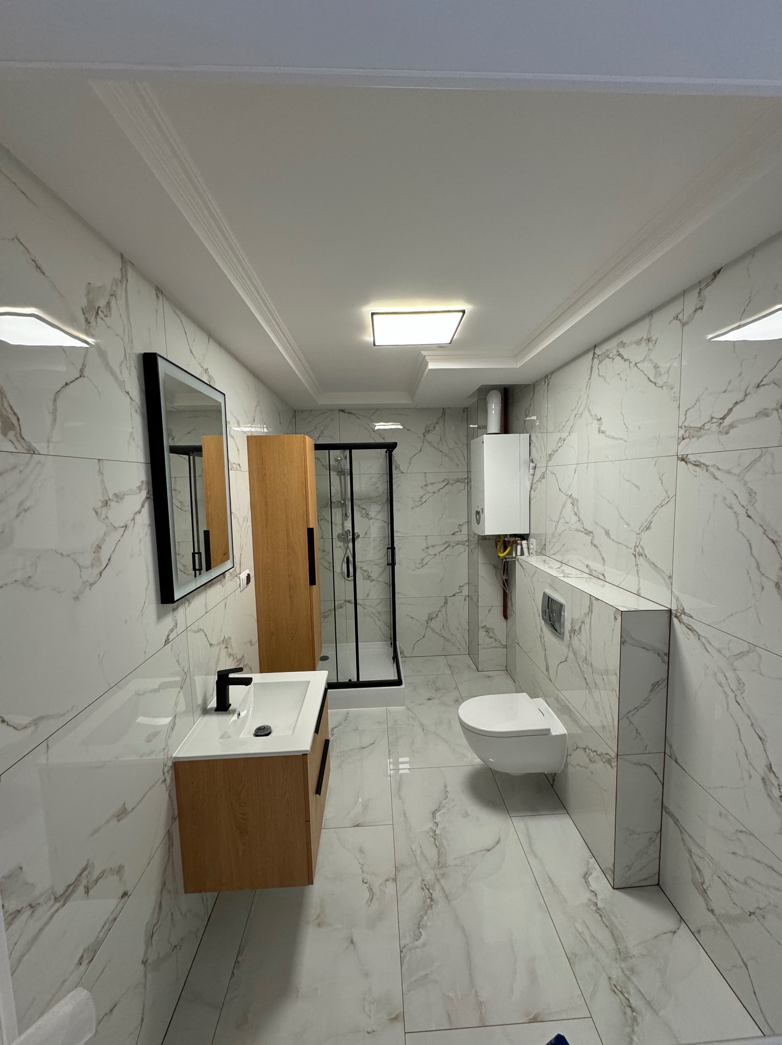 Nowoczesna łazienka z marmurowymi płytkami, kabiną prysznicową z czarnymi ramami, drewnianą szafką pod umywalką z czarną armaturą i podwieszaną toaletą.