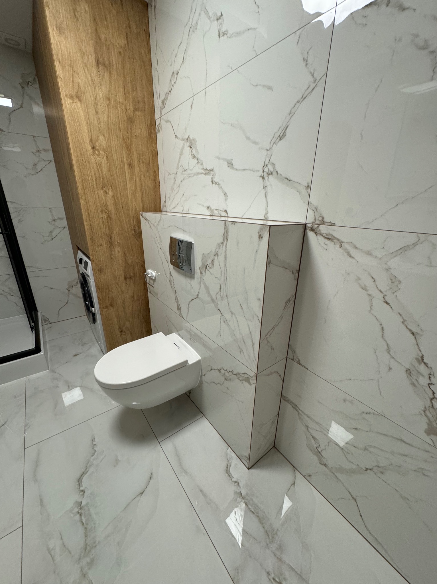 Nowoczesna łazienka z podwieszaną toaletą, płytkami imitującymi marmur na ścianach i podłodze, oraz zabudową z drewnopodobnej płyty, widoczna pralka i kabina prysznicowa.