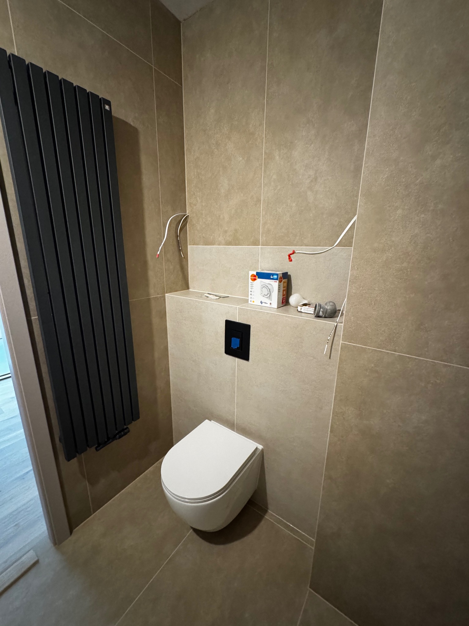 Łazienka w trakcie wykańczania: beżowe płytki, wisząca toaleta, czarny grzejnik żeberkowy, elementy instalacji i pudełko armatury na półce.