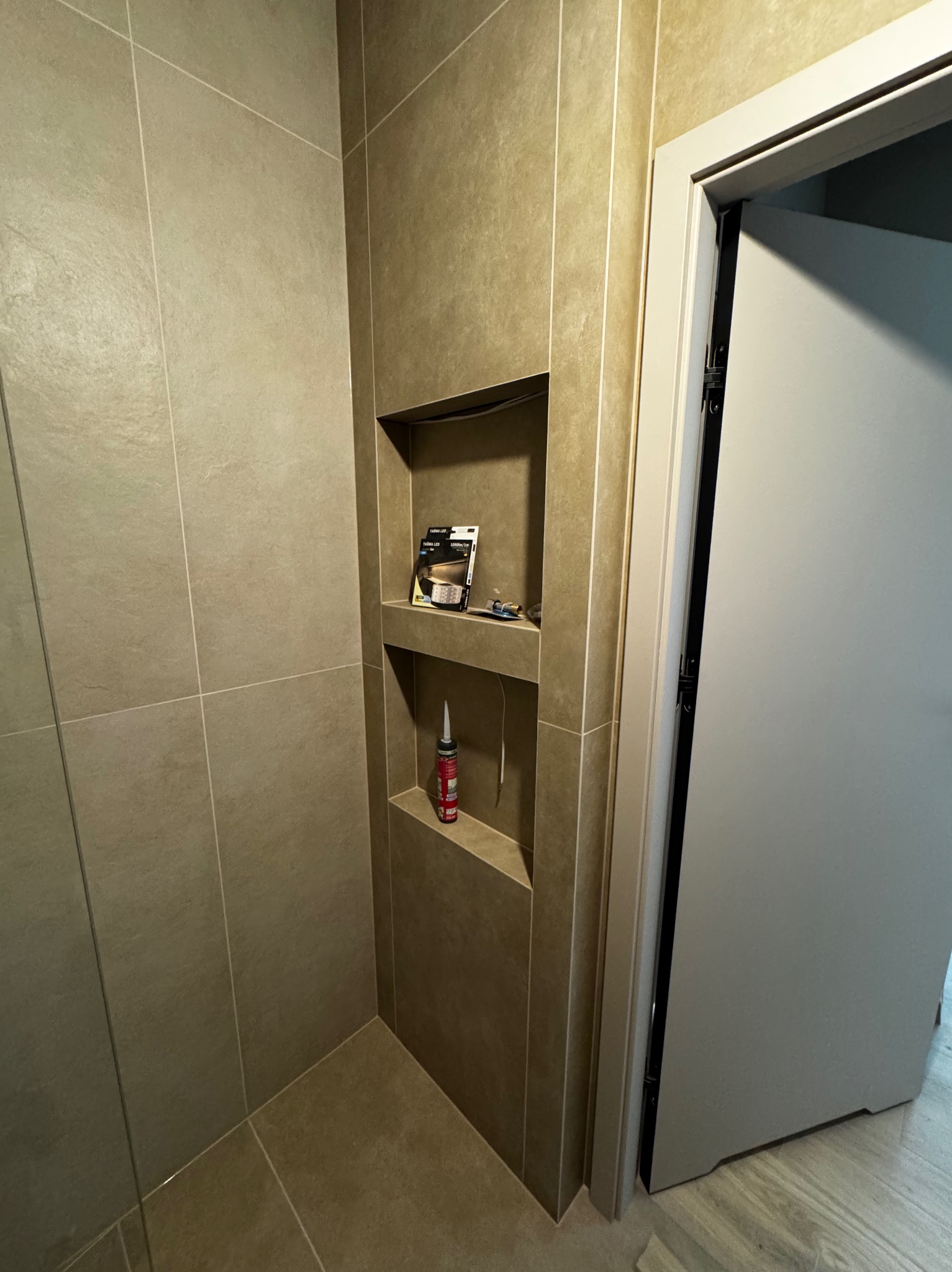 Beżowa łazienka z wnęką wyłożoną płytkami, w której znajdują się pudełka i silikon. Widoczny fragment drzwi. Ukończone wykończenie łazienki.