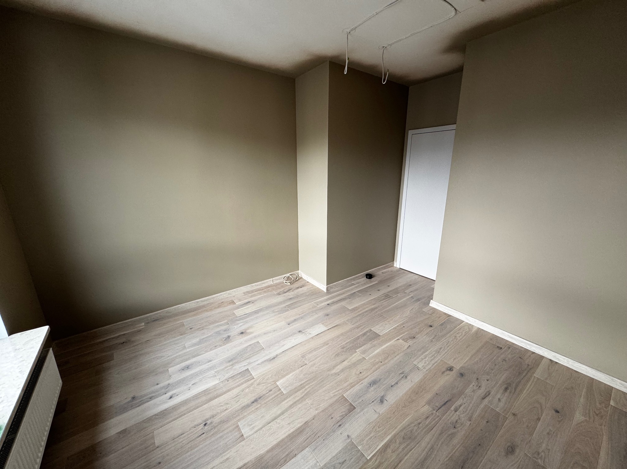 Wnętrze pokoju po malowaniu ścian na beżowo-zielony odcień i ułożeniu jasnej, drewnianej podłogi. Widoczne białe listwy przypodłogowe i wystające kable.