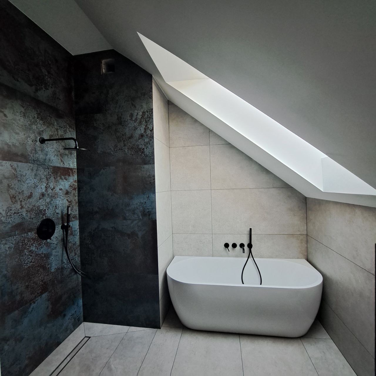 Nowoczesna łazienka na poddaszu z kabiną prysznicową wykończoną płytkami imitującymi rdzę i białą wanną wolnostojącą pod oknem dachowym.