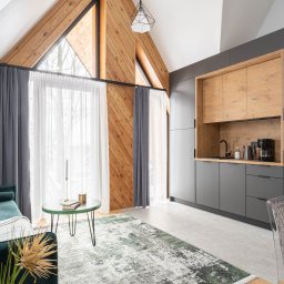 Przedsiębiorstwo Produkcyjno Handlowe ESKA - Nowoczesna kuchnia w szarościach i drewnie w Przytkowicach. Zabudowa kuchenna pod skosem dachu, drewniane wykończenie ściany, okno trójkątne, dywan w odcieniach zieleni.