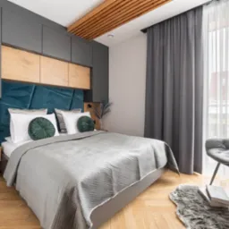 Stylowa sypialnia z szarą zabudową meblową nad łóżkiem i drewnianymi akcentami. Błękitny zagłówek, szare zasłony, drewniany sufit lamelowy.