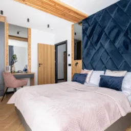 Elegancka sypialnia z łóżkiem, toaletką i szafą wnękową. Drewniane elementy dekoracyjne i niebieski zagłówek dodają wnętrzu charakteru. Widoczny parkiet i oświetlenie.