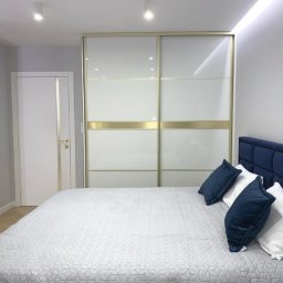 Przedsiębiorstwo Produkcyjno Handlowe ESKA - Szafa wnękowa z przesuwnymi drzwiami w sypialni, białe fronty z poziomymi złotymi akcentami. Widoczne łóżko z niebieskim zagłówkiem i biała kołdra.