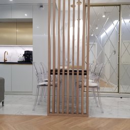 Przedsiębiorstwo Produkcyjno Handlowe ESKA - Nowoczesna kuchnia z białymi i złotymi szafkami, marmurowym blatem, szklanym panelem i drewnianą ścianką działową z transparentnymi krzesłami.