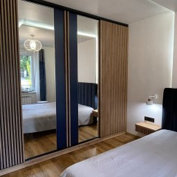 Przedsiębiorstwo Produkcyjno Handlowe ESKA - Nowoczesna szafa wnękowa z lustrzanymi drzwiami przesuwnymi i drewnianymi lamelami, wkomponowana w aranżację sypialni z parkietem i oświetleniem.