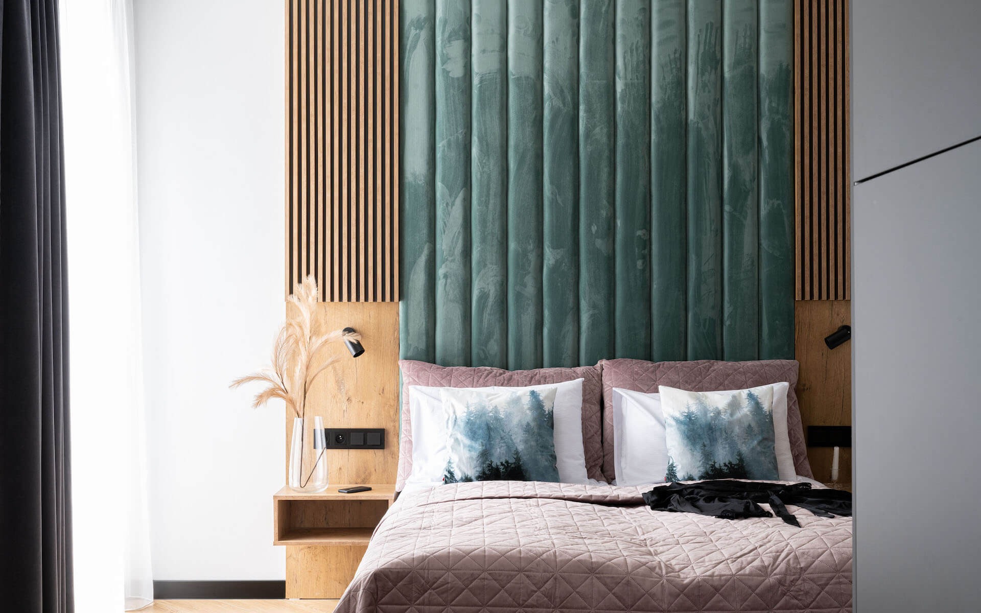 Nowoczesna sypialnia z tapicerowanym, zielonym panelem ściennym i drewnianymi lamelami, uzupełniona o szafkę nocną z drewna i lampkę.