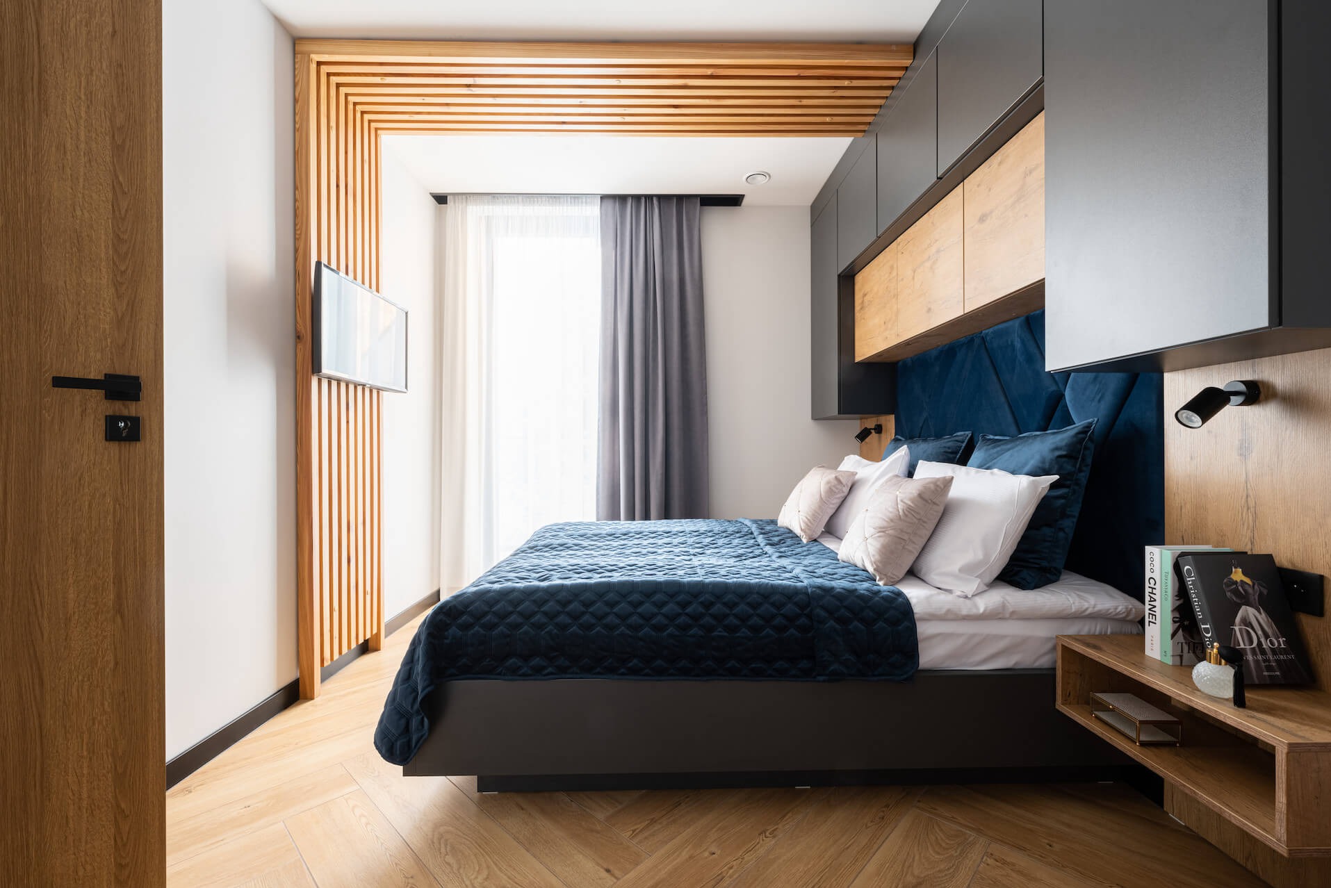 Nowoczesna sypialnia z drewnianą zabudową nad łóżkiem, szafkami i telewizorem. Niebieskie obicie łóżka i parkiet w jodełkę. Stylowe oświetlenie i dodatki.
