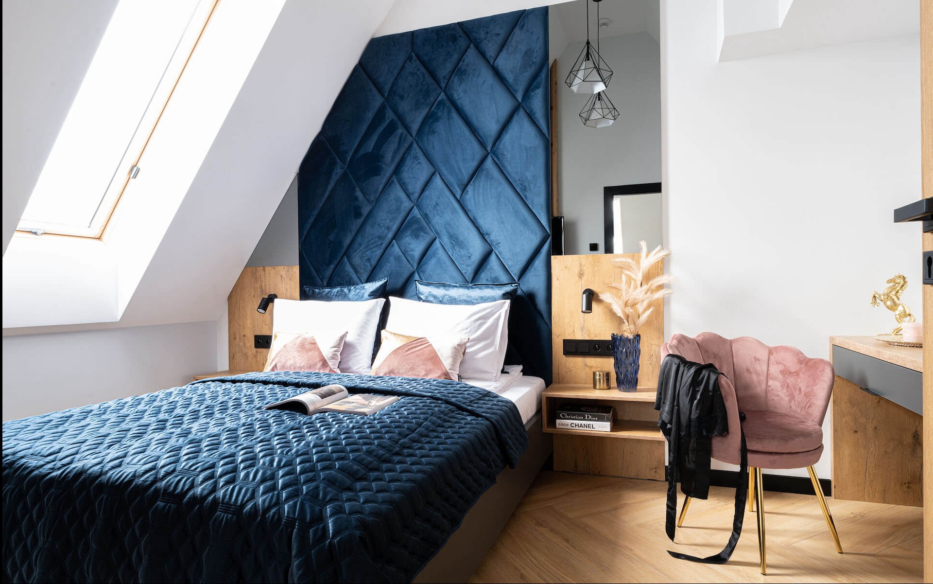Sypialnia na poddaszu z łóżkiem z pikowanym, granatowym zagłówkiem, drewnianymi szafkami nocnymi i oknem dachowym. Krzesło tapicerowane w stylu glamour.