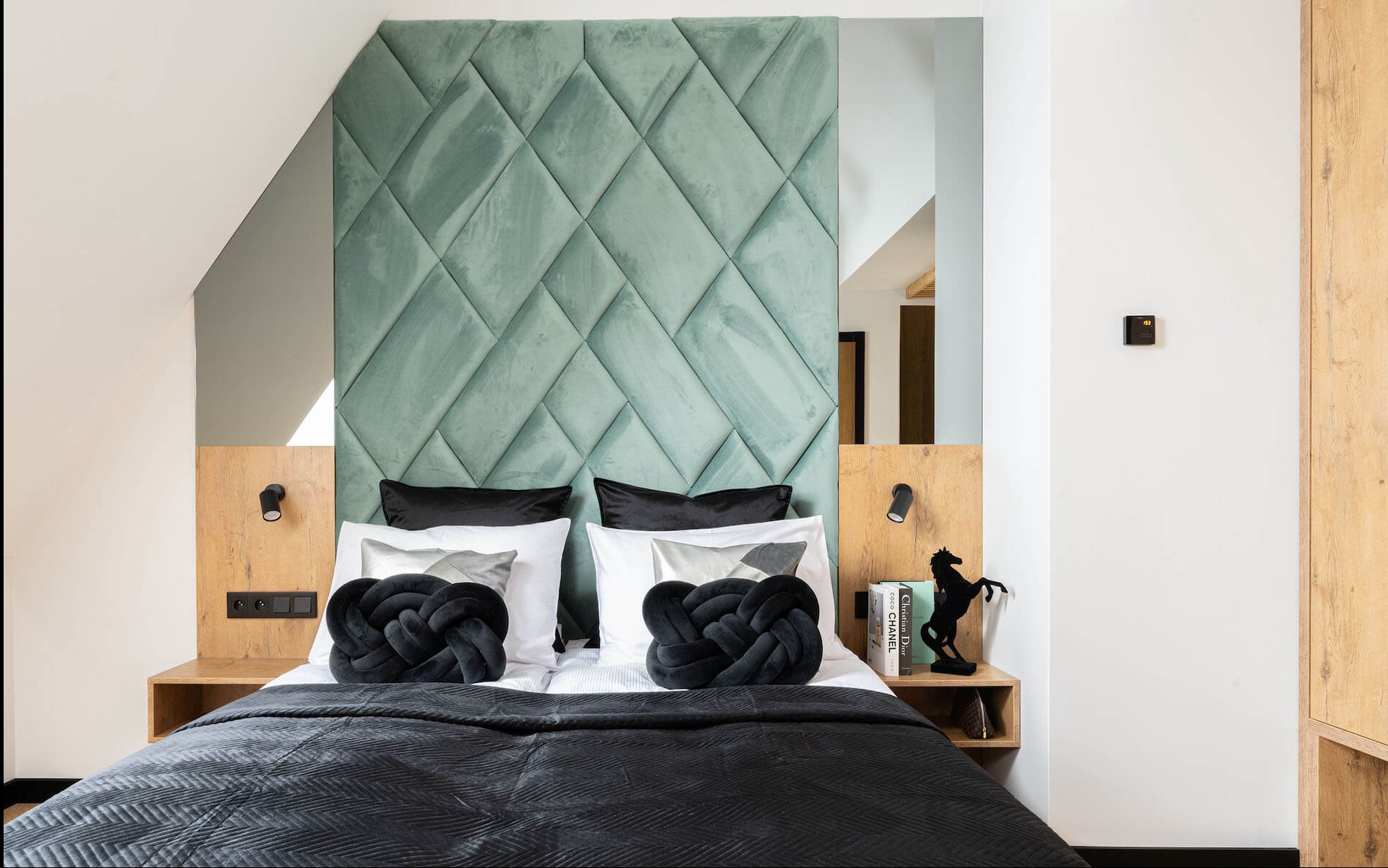 Sypialnia z pikowanym, miętowym zagłówkiem, drewnianymi szafkami nocnymi i czarnymi poduszkami w kształcie węzłów. Nowoczesny design z oświetleniem punktowym.