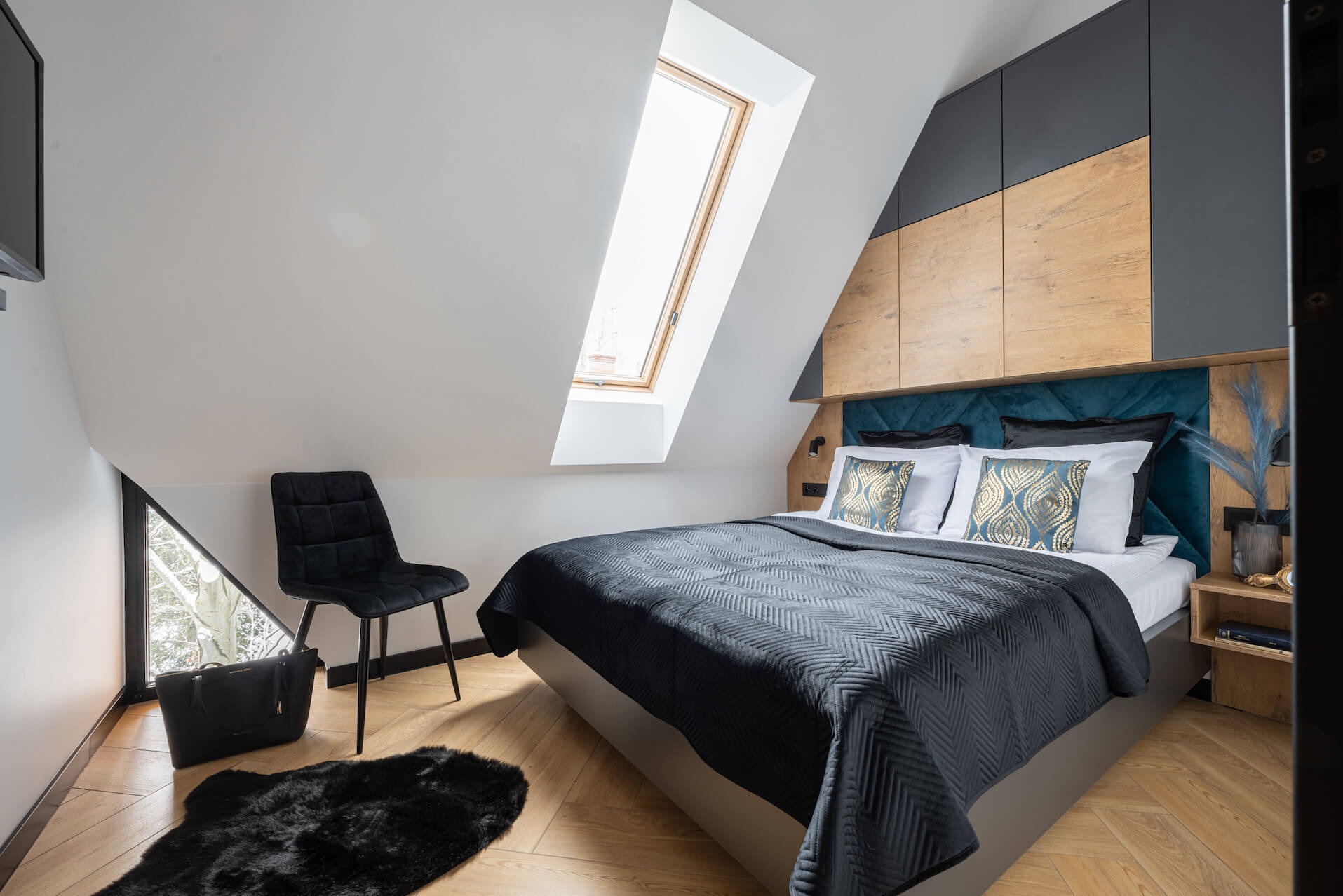 Stylowa sypialnia na poddaszu z łóżkiem, szafkami w zabudowie i oknem dachowym. Uwagę przykuwa ciemna kolorystyka i skośny sufit. Na podłodze parkiet i czarny dywan.