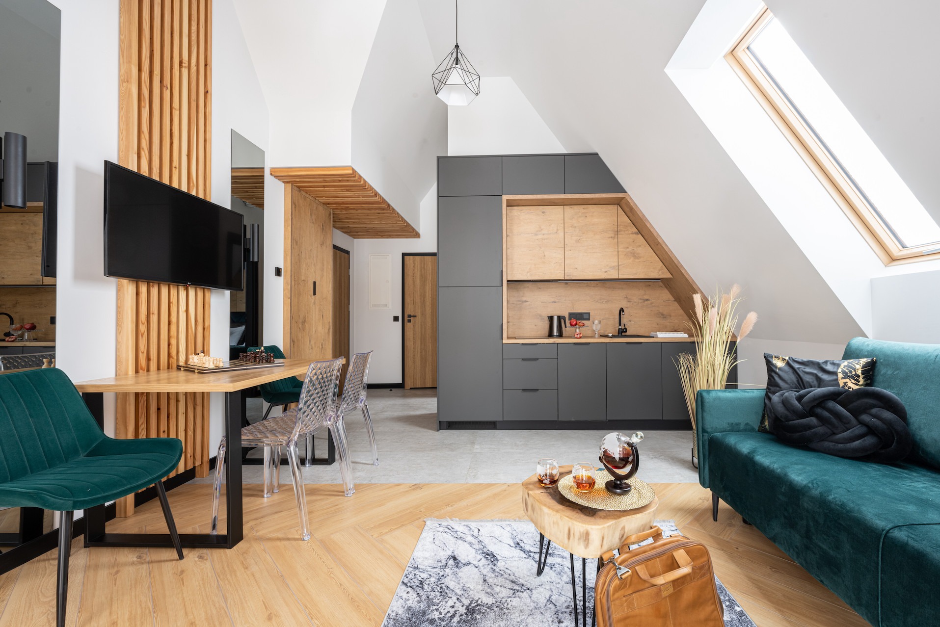 Nowoczesne wnętrze ze skośnym sufitem i oknem dachowym. Kuchnia w szarościach i drewnie, stół z szachami, zielona sofa i drewniane akcenty dekoracyjne.