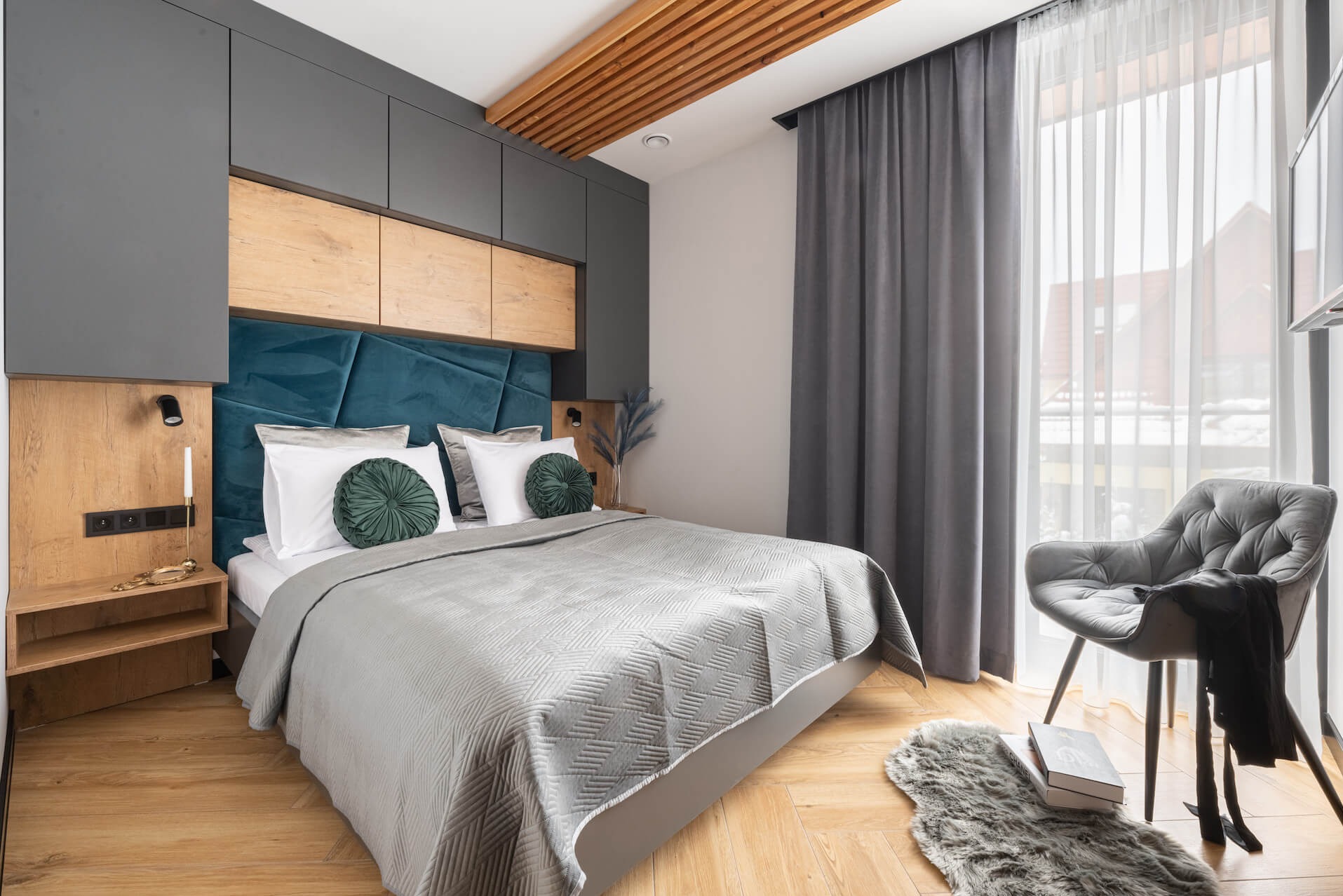 Stylowa sypialnia z szarą zabudową meblową nad łóżkiem i drewnianymi akcentami. Błękitny zagłówek, szare zasłony, drewniany sufit lamelowy.