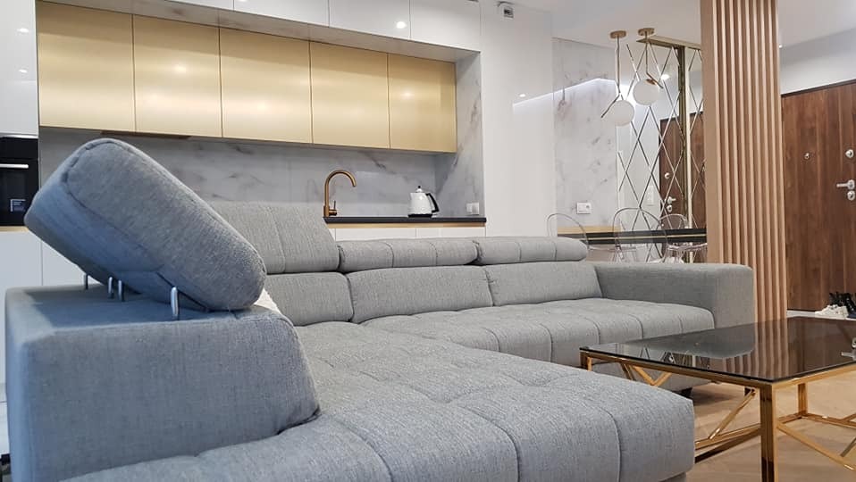 Szary narożnik z regulowanymi zagłówkami w nowoczesnym salonie z aneksem kuchennym. Złote szafki kuchenne i armatura, marmurowy backsplash.