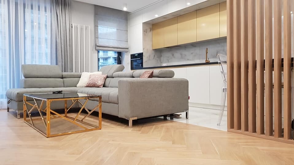 Nowoczesne wnętrze z szarą sofą narożną, złotym stolikiem kawowym i zabudową kuchenną z elementami złota, parkiet w jodełkę, drewniana ażurowa ścianka działowa.