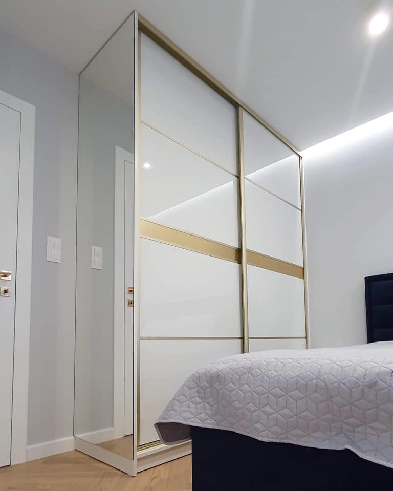 Nowoczesna szafa wnękowa z lustrzanymi drzwiami przesuwnymi i złotymi akcentami, w jasnym wnętrzu sypialni z widocznym fragmentem łóżka.