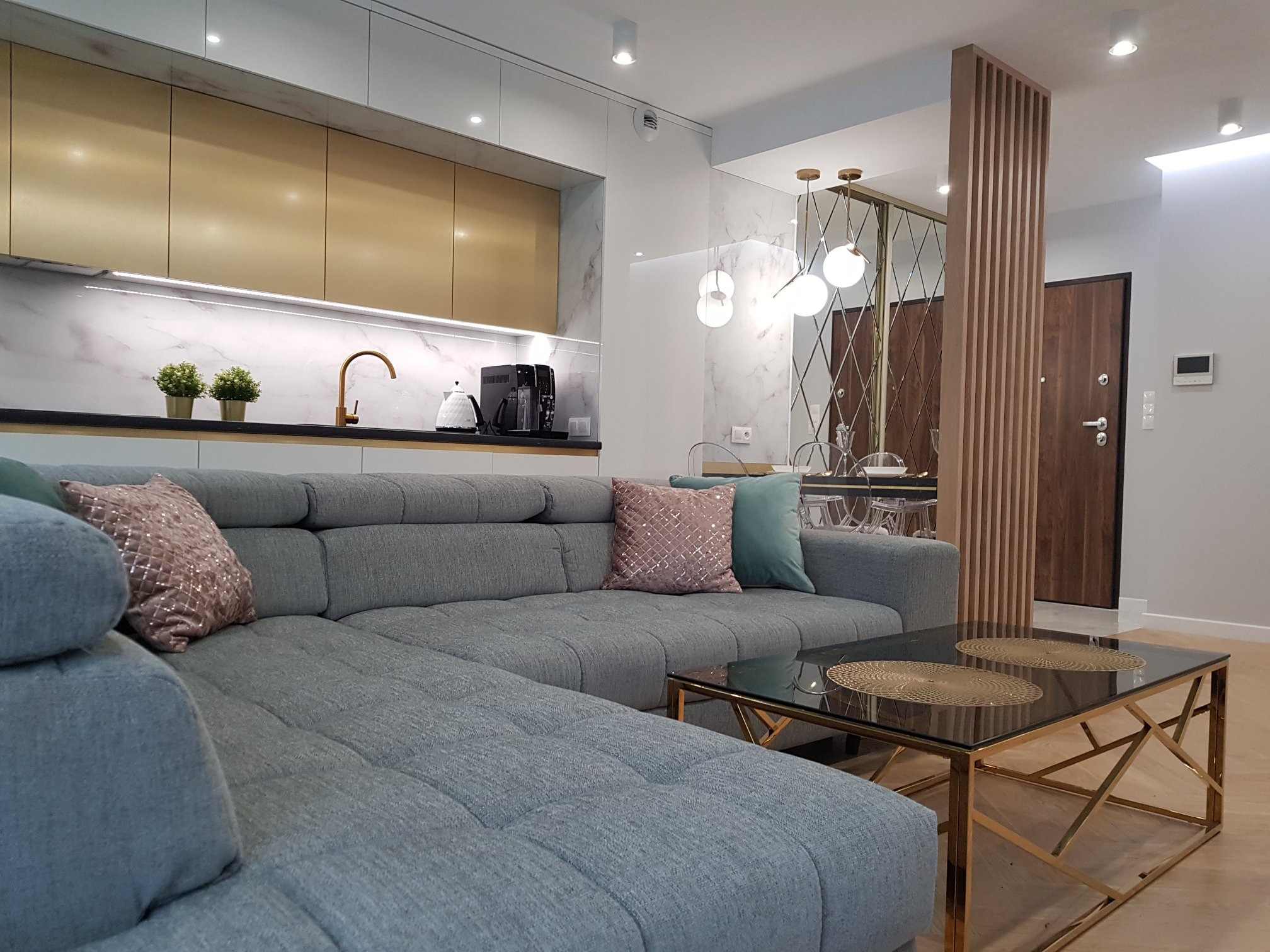 Nowoczesne wnętrze: szara sofa narożna, złota kuchnia z marmurowym backsplashiem, szklany stolik kawowy ze złotą ramą i dekoracyjna ścianka działowa.