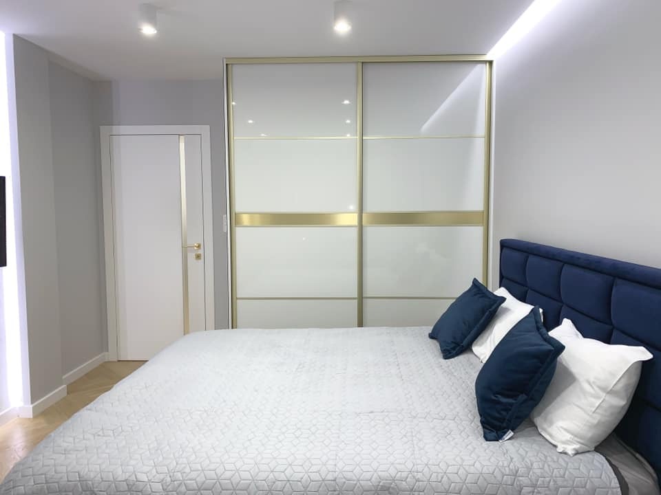 Szafa wnękowa z przesuwnymi drzwiami w sypialni, białe fronty z poziomymi złotymi akcentami. Widoczne łóżko z niebieskim zagłówkiem i biała kołdra.
