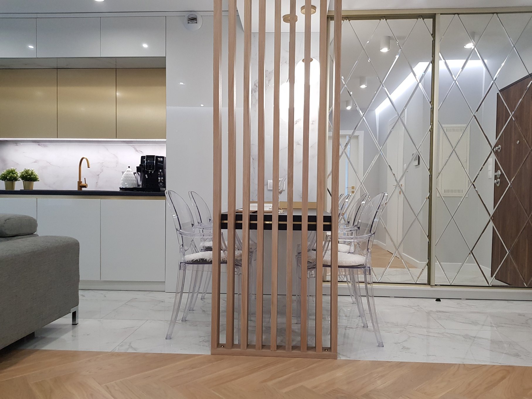 Nowoczesna kuchnia z białymi i złotymi szafkami, marmurowym blatem, szklanym panelem i drewnianą ścianką działową z transparentnymi krzesłami.
