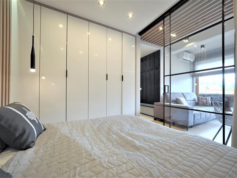 Biała szafa wnękowa z połyskiem w sypialni, widok z łóżka, czarne uchwyty, szklana ściana oddzielająca od salonu, nowoczesny design, minimalistyczny styl.