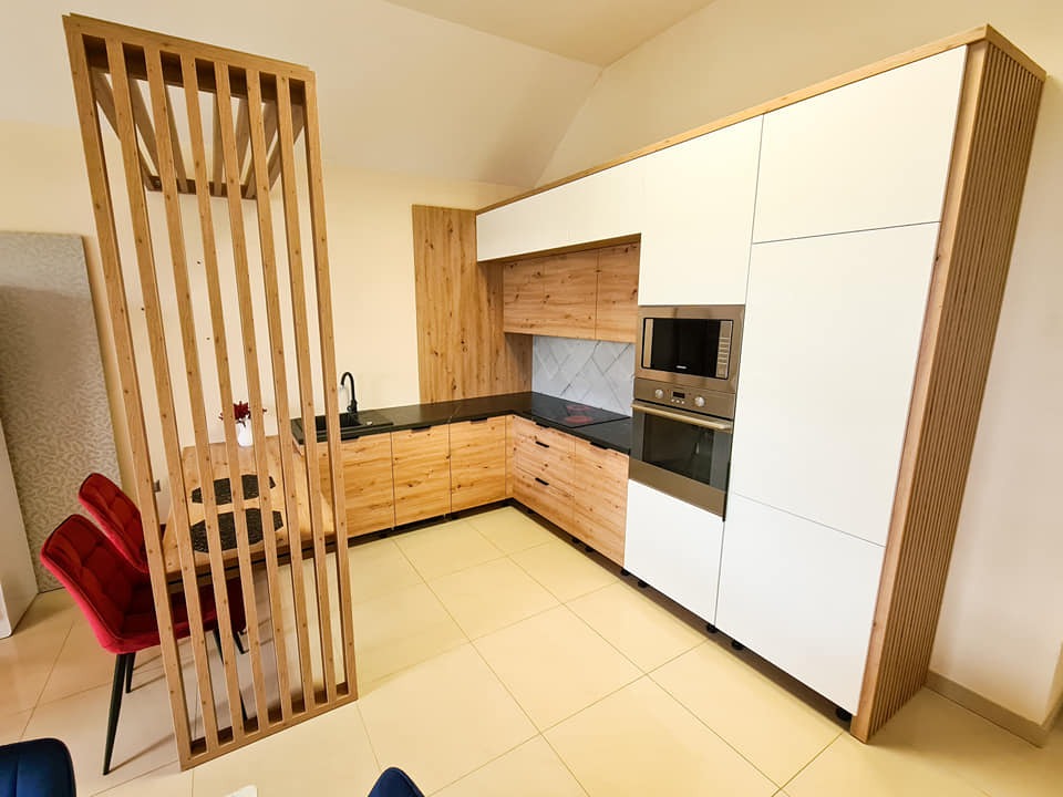 Jasna kuchnia na wymiar z drewnianymi frontami i białymi szafkami, oddzielona ażurową ścianką od jadalni z czerwonymi krzesłami. Nowoczesne wykończenie.