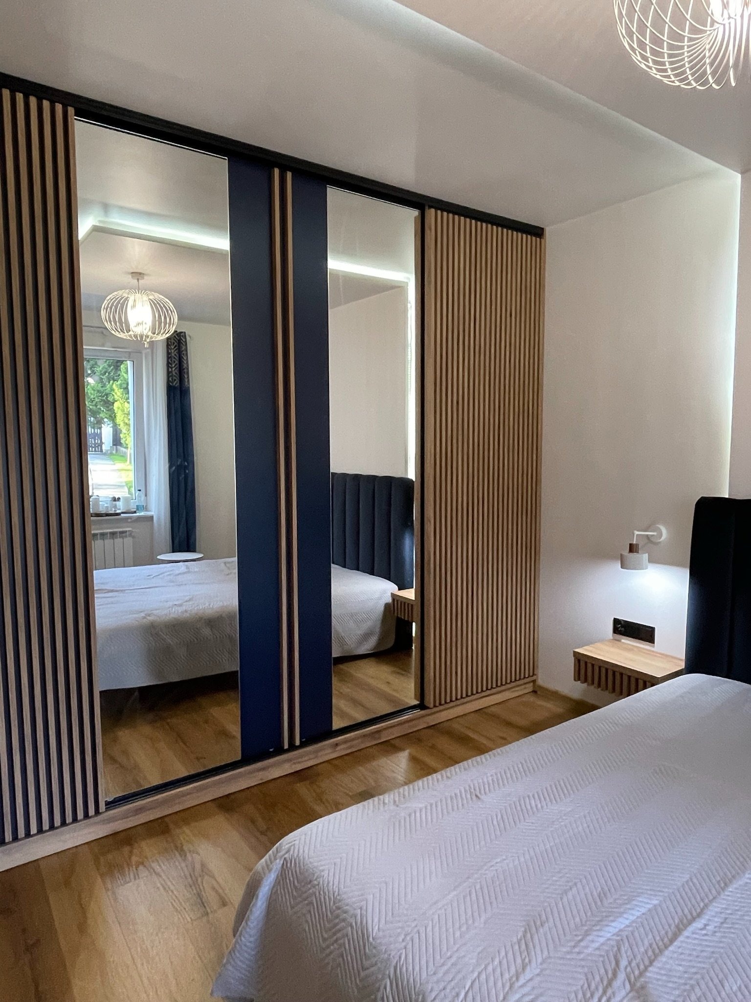 Nowoczesna szafa wnękowa z lustrzanymi drzwiami przesuwnymi i drewnianymi lamelami, wkomponowana w aranżację sypialni z parkietem i oświetleniem.