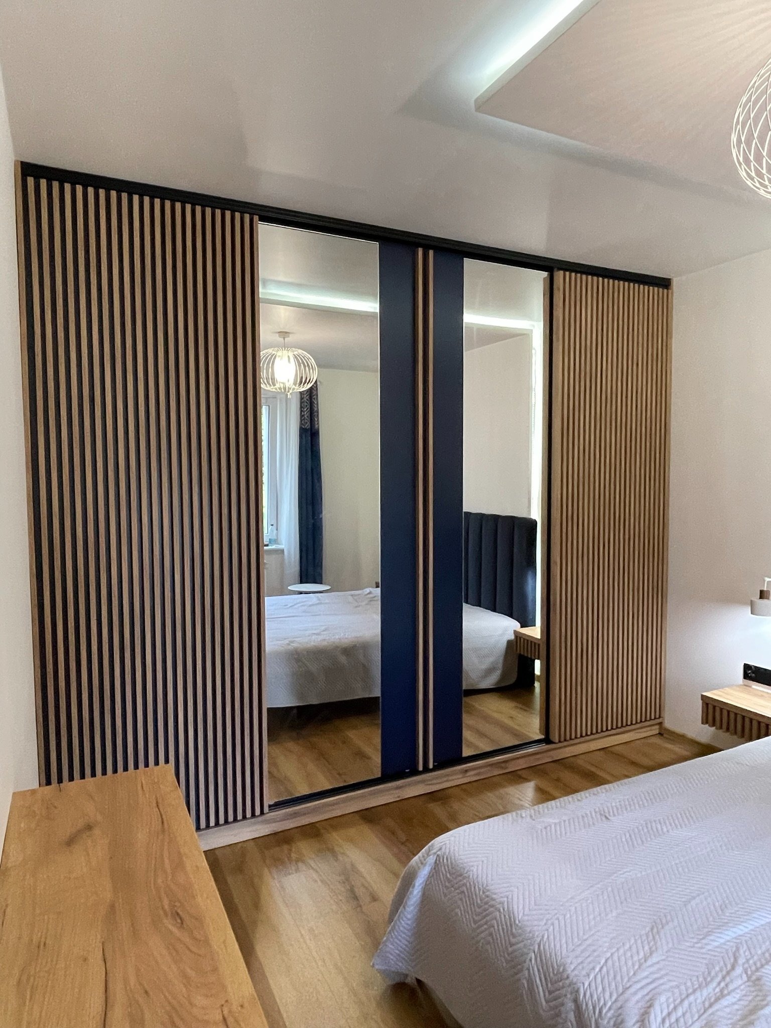 Nowoczesna szafa wnękowa z lamelami drewnianymi i lustrzanymi przesuwnymi drzwiami, odbijającymi wnętrze sypialni. Minimalistyczny design, ciepłe oświetlenie.