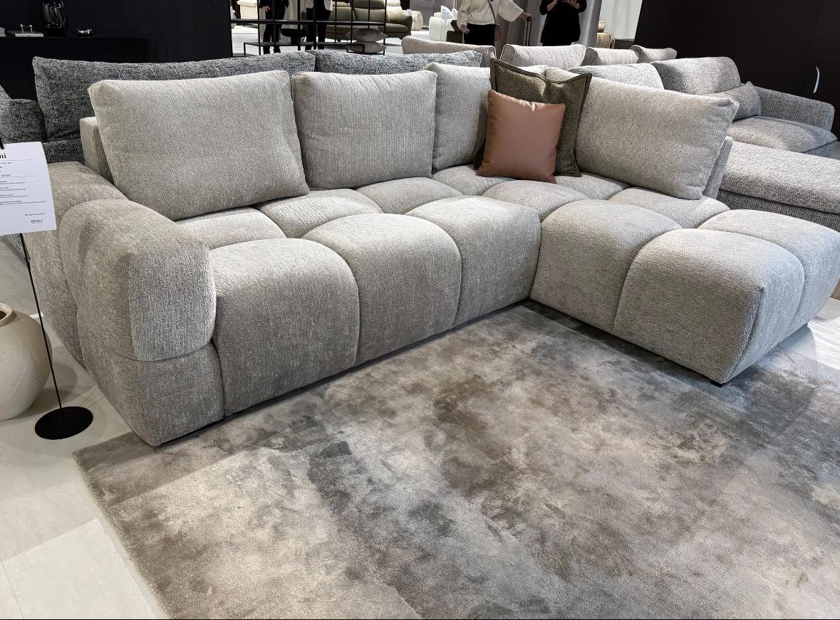 Szary, modułowy narożnik z miękkimi poduchami i brązową poduszką, ustawiony na szarym dywanie. Widoczny fragment ekspozycji meblowej.