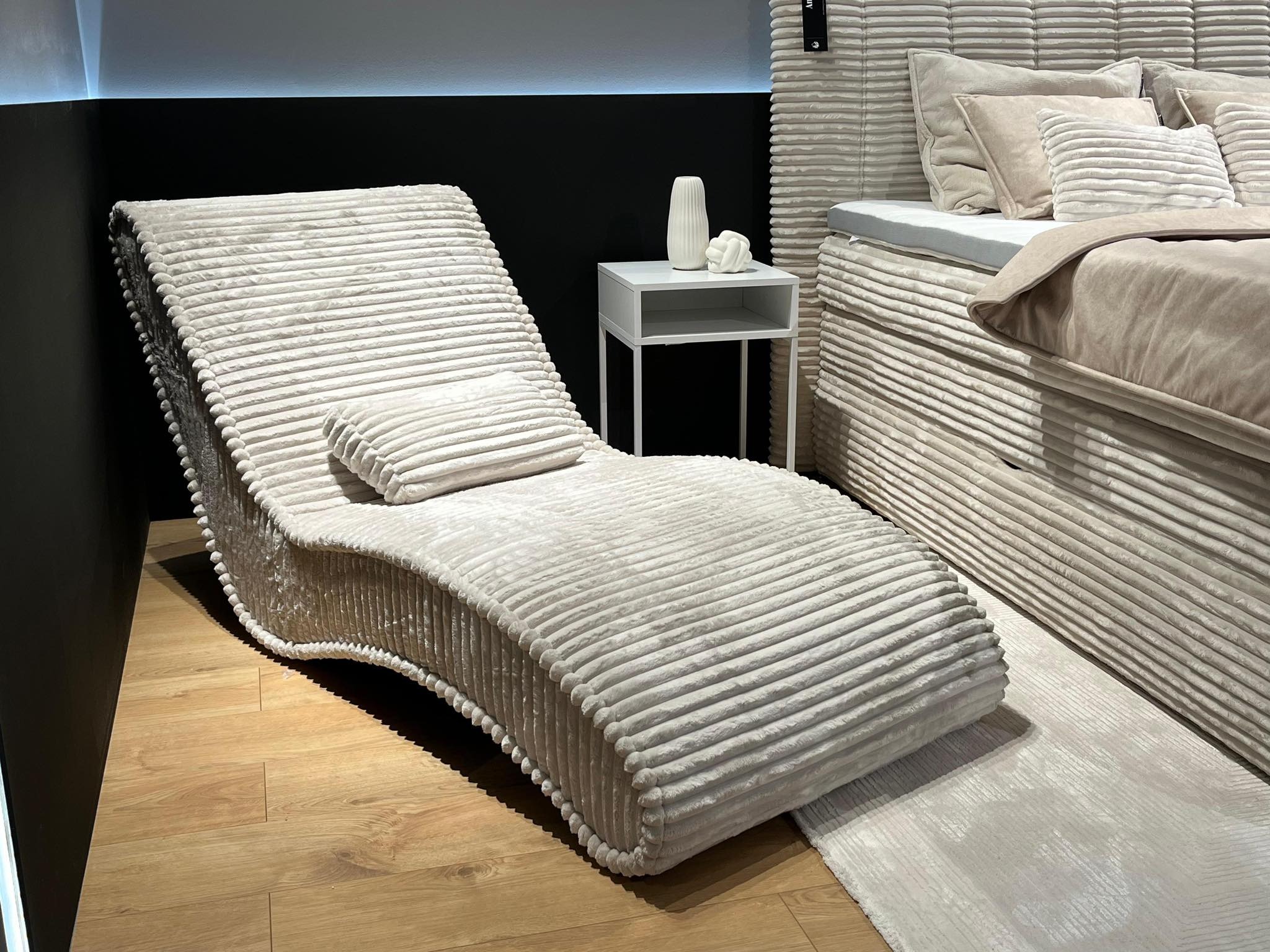 Beżowy, prążkowany leżak obok łóżka z wezgłowiem w identycznej stylistyce. Na stoliku nocnym białe wazony. Całość w nowoczesnym wnętrzu.