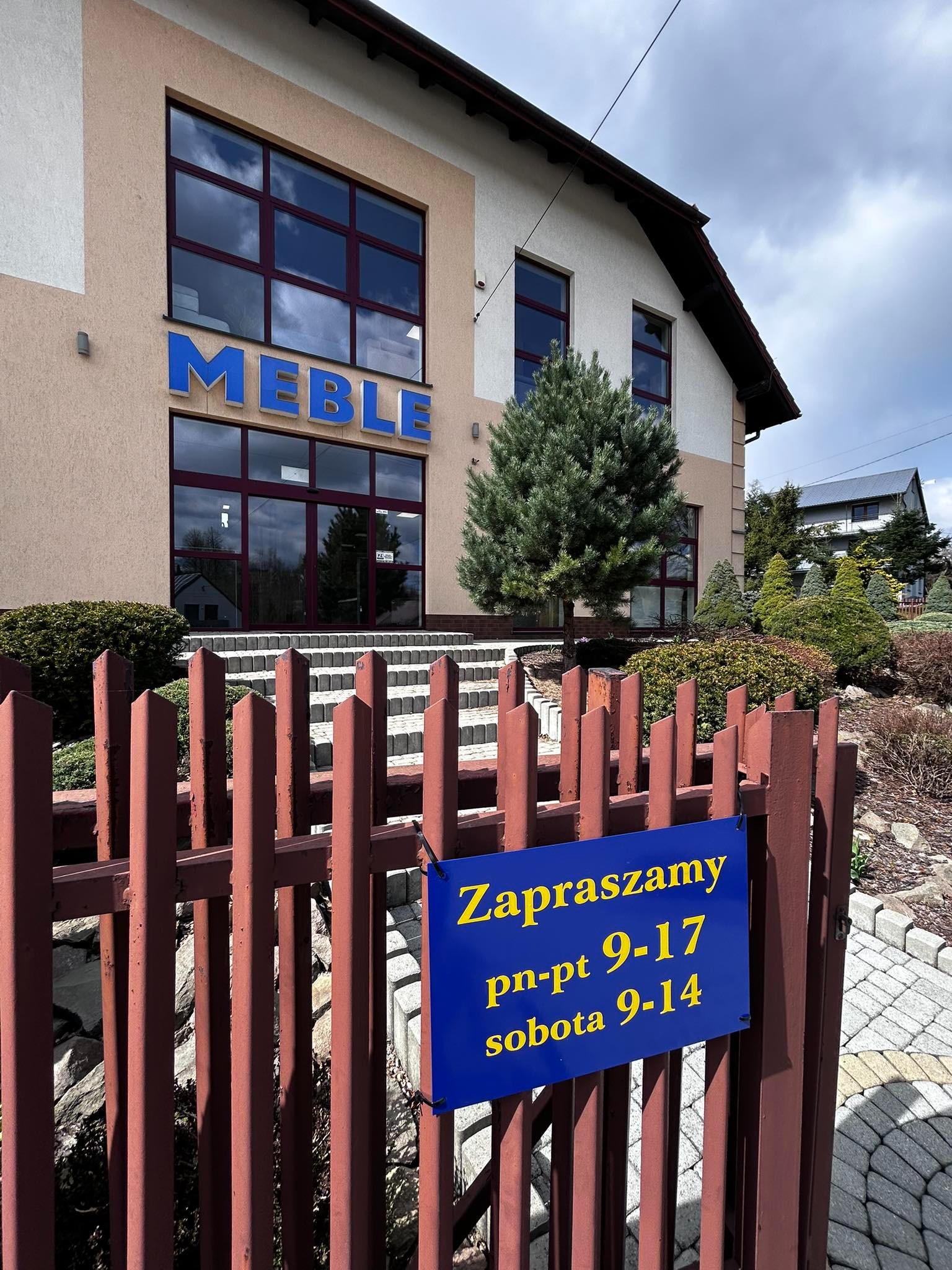 Budynek z napisem 'MEBLE' nad wejściem, widoczny przez brązowy płot z niebieską tabliczką z godzinami otwarcia. Ujęcie z zewnątrz, w tle zieleń.
