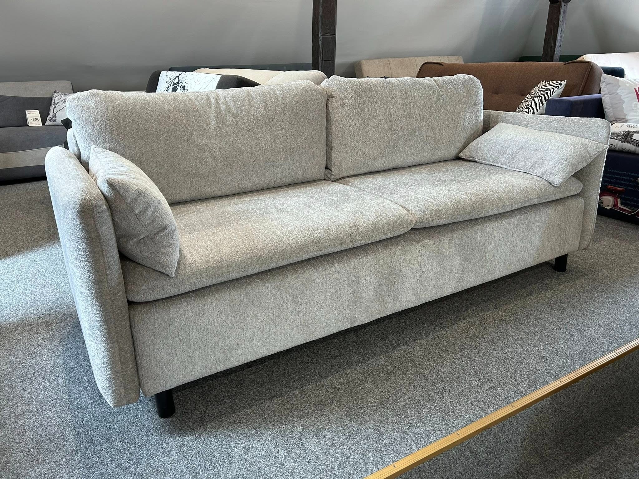 Jasnoszara, dwuosobowa sofa z poduszkami na tle innych mebli w salonie. Minimalistyczny design, proste linie, na niskich nóżkach. Widoczny fragment podłogi.