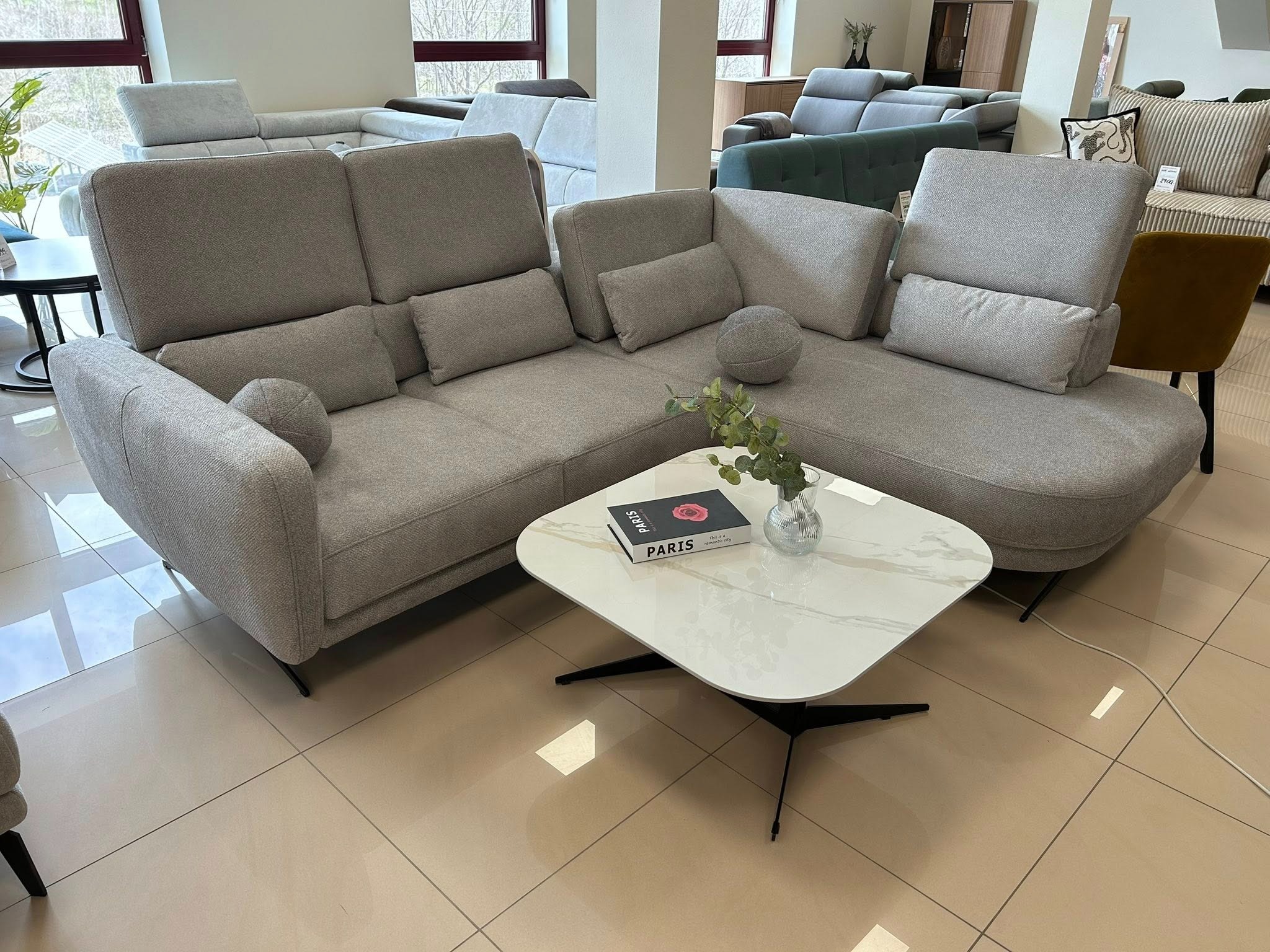 Jasnoszara sofa narożna z poduszkami i stolikiem kawowym na beżowych płytkach. W tle inne sofy i okna.