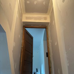 Łukasz Pierz - Wnętrze w trakcie remontu: ściany i sufit po szpachlowaniu, widoczne punkty po łataniu, sufit w trakcie przygotowania, wisząca żarówka, otwarta drewniana rama drzwiowa prowadząca do jasnego...