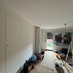 Łukasz Pierz - Pokój w trakcie remontu: pomalowane ściany na biało, zabezpieczona podłoga, drabina, narzędzia i materiały malarskie widoczne w pomieszczeniu.