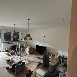 Łukasz Pierz - Pokój w trakcie remontu: drabina, narzędzia, lampy wiszące i świeżo pomalowane ściany z widocznymi otworami na kable. Podłoga parkietowa częściowo zakryta materiałami zabezpieczającymi.