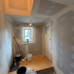 Łukasz Pierz - Wnętrze w trakcie remontu: ściany częściowo pokryte szpachlą, widoczne narzędzia i wiaderka z zaprawą, drewniana podłoga zabezpieczona przed zabrudzeniem, okno z widokiem na zieleń.