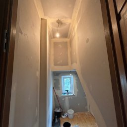 Łukasz Pierz - Korytarz w trakcie remontu, ściany częściowo pokryte masą szpachlową, widoczne narzędzia i wiaderka, surowe drewniane podłogi w dolnej części korytarza, okno w tle.