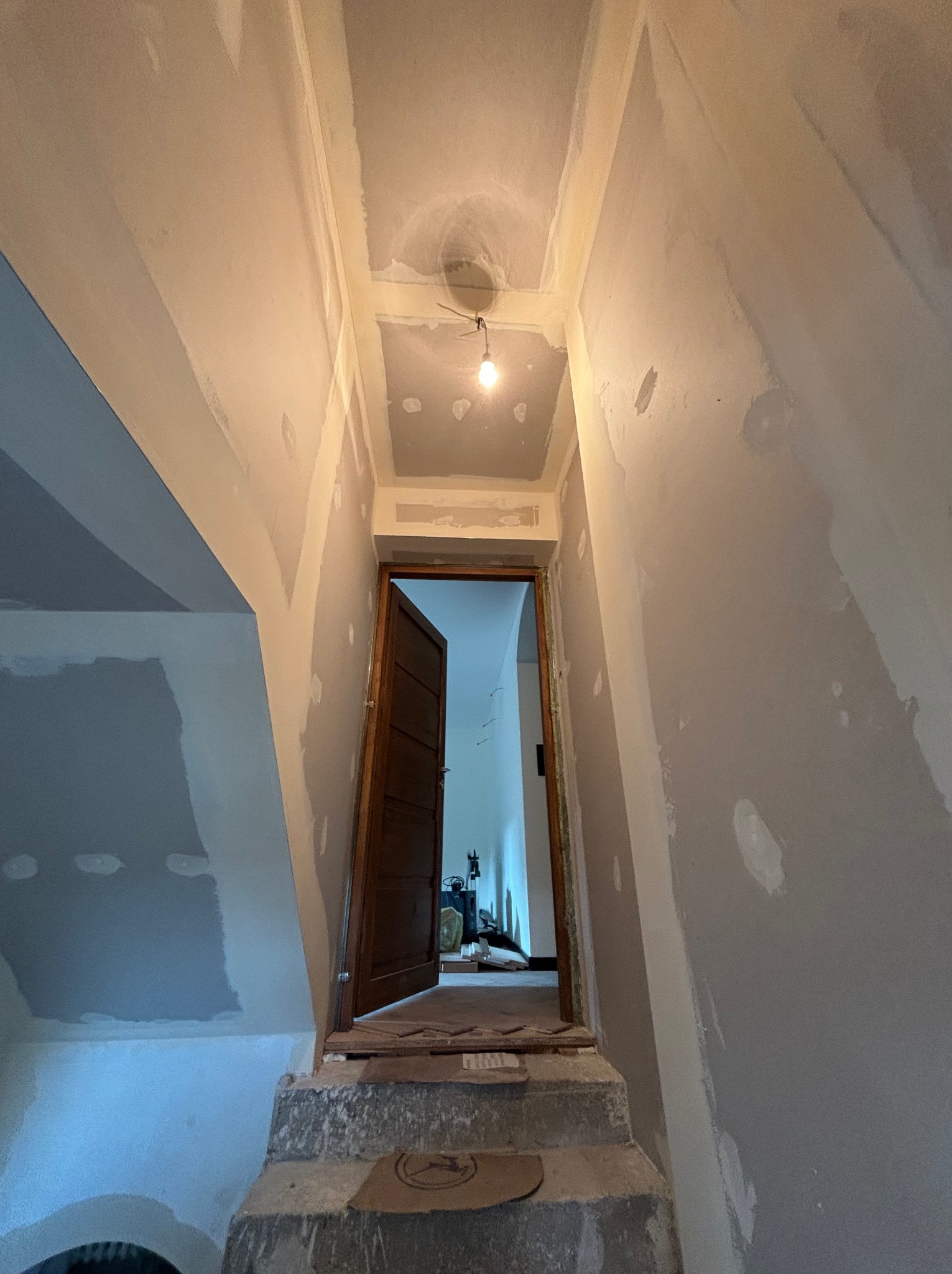 Wnętrze klatki schodowej w trakcie prac wykończeniowych, ściany pokryte warstwą szpachli, widoczne ślady po łataniu ubytków, otwarte drzwi prowadzące do dalszej części domu, surowa żarówka...