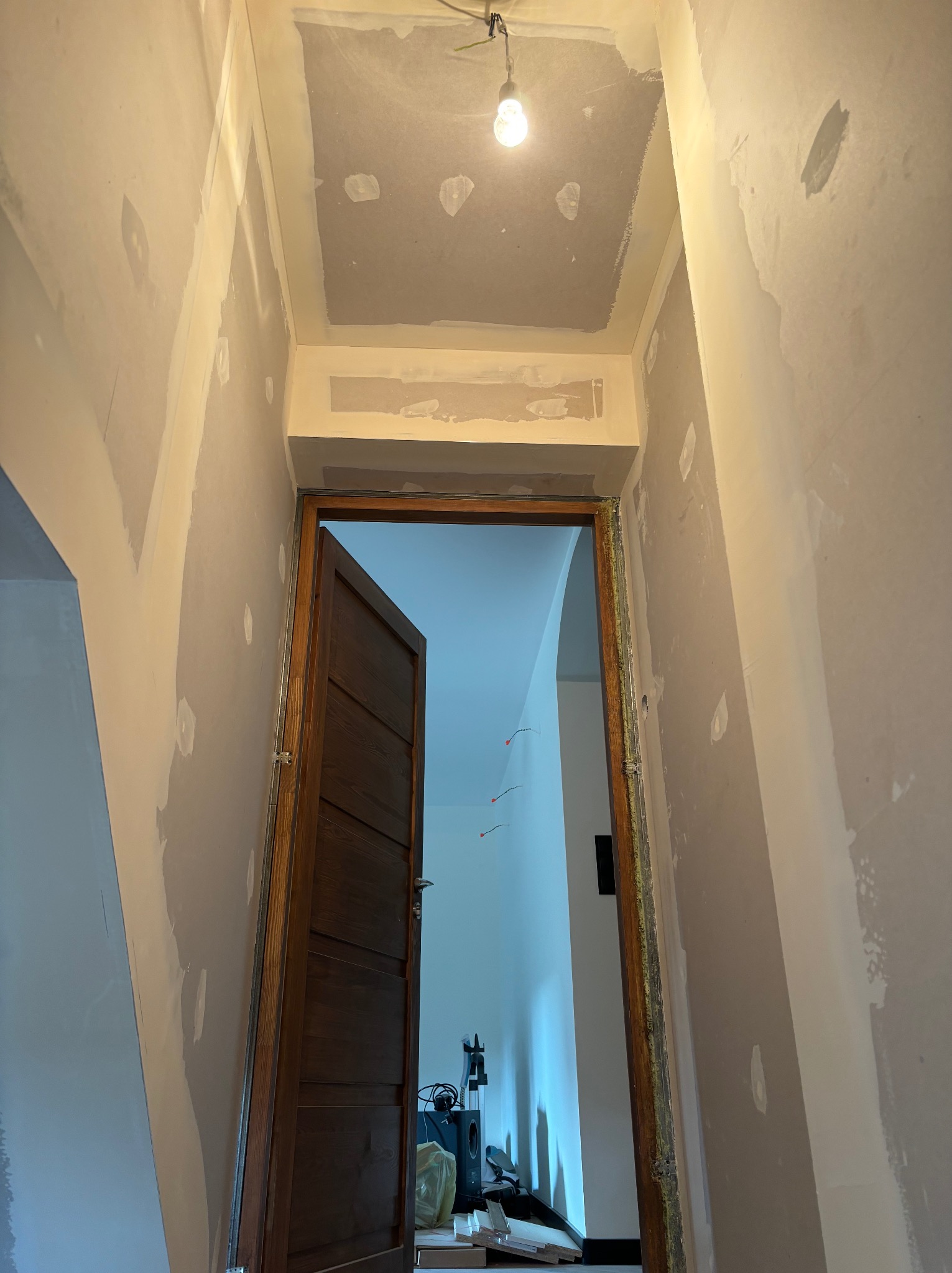 Wnętrze w trakcie remontu: ściany i sufit po szpachlowaniu, widoczne punkty po łataniu, sufit w trakcie przygotowania, wisząca żarówka, otwarta drewniana rama drzwiowa prowadząca do jasnego...