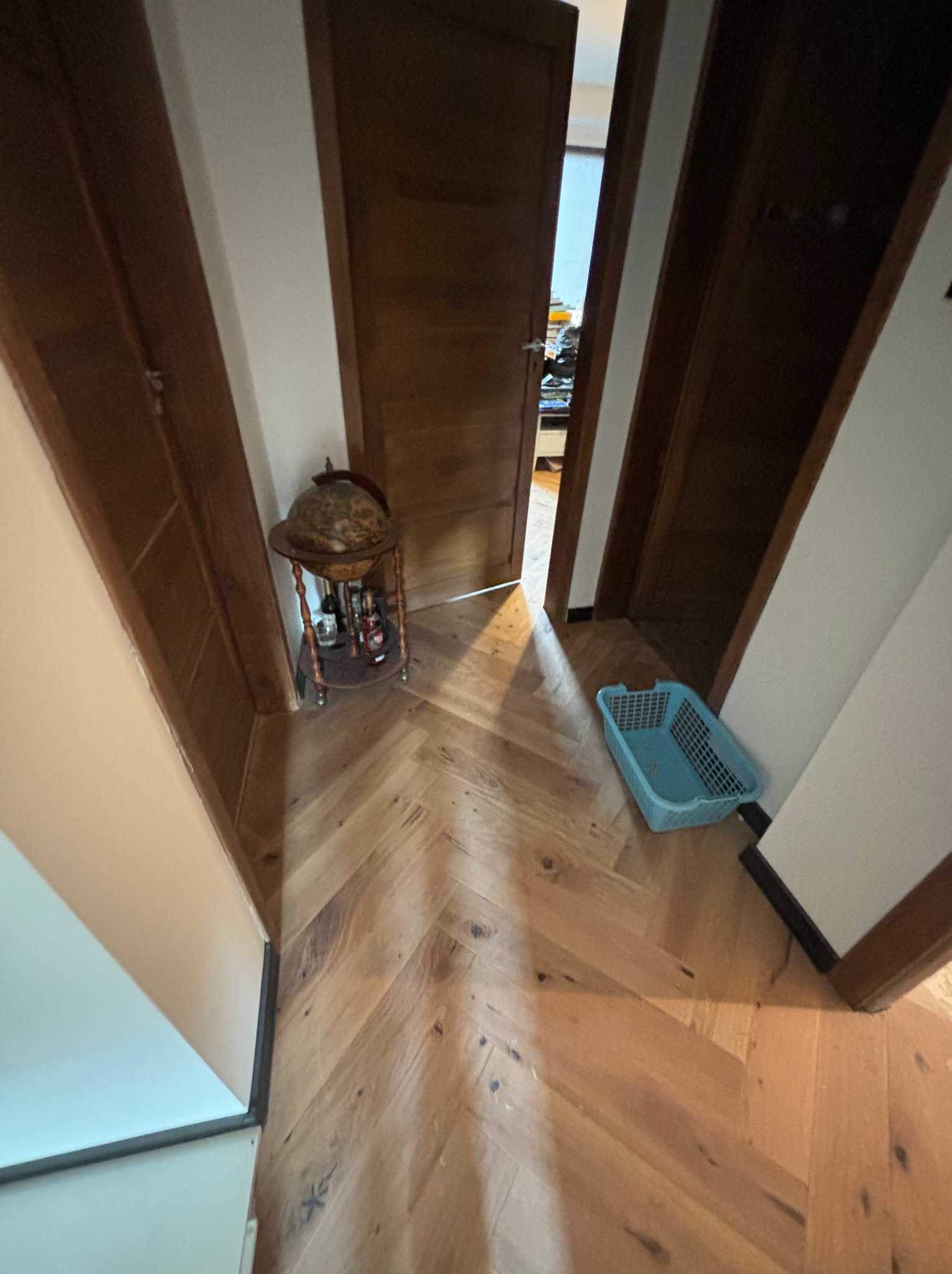 Korytarz z parkietem ułożonym w jodełkę, widoczne drzwi i szafka na alkohol z globusem na tle otwartych drzwi.